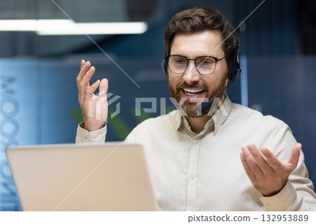 Close-up photo of a young smiling man in a headset sitting in the office at a table in front of a laptop and talking on a video call. Close-up photo of a young smiling man in a headset sitting in the office at a table in front of a laptop and talking on a video call. 132953889