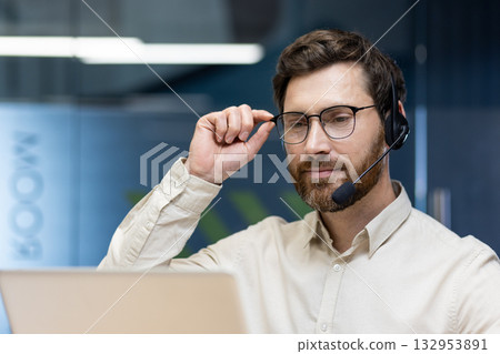 Close-up photo of a young serious man in a headset sitting in the office at a laptop, looking at the screen and holding his glasses with his hand. Close-up photo of a young serious man in a headset sitting in the office at a laptop, looking at the screen and holding his glasses with his hand. 132953891