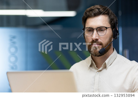 Close-up photo of a young serious and confident office worker in glasses and a headset working with a laptop. 132953894