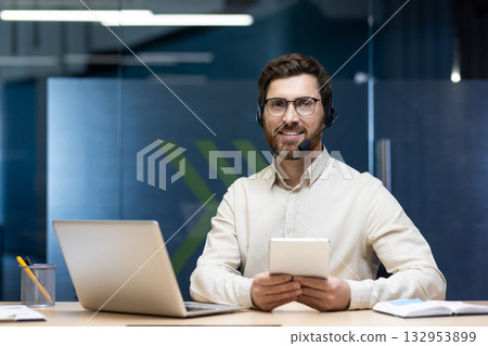 Portrait of a young businessman in a headset sitting at a workplace with a laptop, holding a tablet in his hands and looking at the camera with a smile. 132953899