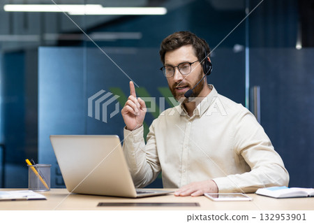 Serious young man sitting in the office at the desk in a headset and talking on a video call on a laptop, explaining, pointing. 132953901