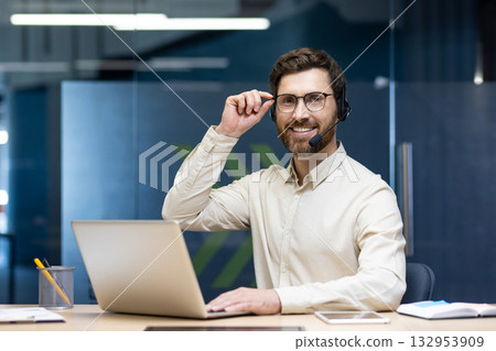 Portrait of a young smiling man wearing a headset sitting at a desk in the office, working on a laptop, holding glasses and looking at the camera. Portrait of a young smiling man wearing a headset sitting at a desk in the office, working on a laptop, holding glasses and looking at the camera. 132953909