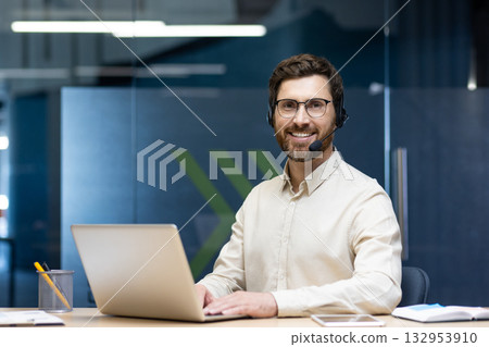 Portrait of a smiling young man in a headset sitting at a desk in the office, using a laptop and looking confidently at the camera. Portrait of a smiling young man in a headset sitting at a desk in the office, using a laptop and looking confidently at the camera. 132953910