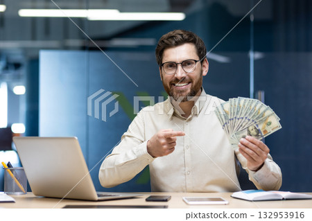 Portrait of a smiling young man sitting in an office at a desk with a laptop, holding a fan of banknotes and pointing at dollars. Portrait of a smiling young man sitting in an office at a desk with a laptop, holding a fan of banknotes and pointing at dollars. 132953916