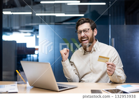 Happy young man sitting in office at desk with laptop, holding credit card in hand and happy showing victory gesture with hand. 132953938