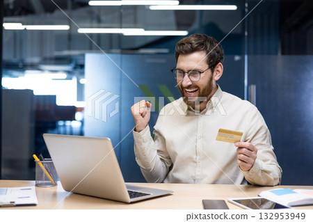 A happy young man is in the office, sitting at the table. is holding a credit card, looking at the laptop screen and showing a victory gesture with his hand. A happy young man is in the office, sitting at the table. is holding a credit card, looking at the laptop screen and showing a victory gesture with his hand. 132953949
