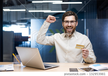 Portrait of a happy young man sitting in the office at a desk with a laptop, holding a credit card in his hands and rejoicing in financial success at the camera, raising his hand up. 132953981