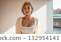 Young woman poses confidently indoors while wearing a cozy cardigan and a fitted top near a sunny window in the late morning 132954141