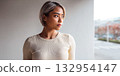 Woman with short hair gazes thoughtfully out of a window in an urban setting during daytime 132954147
