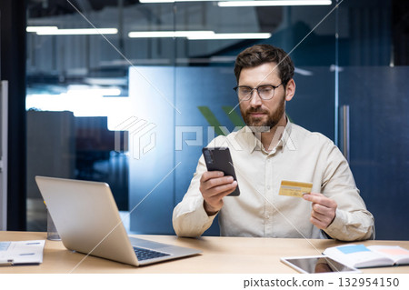 A serious young businessman is sitting in the office at the desk, holding a credit card and using a mobile phone. 132954150