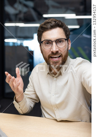 Vertical close-up photo of a young smiling male businessman holding an online meeting in the office, holding a camera. 132954157