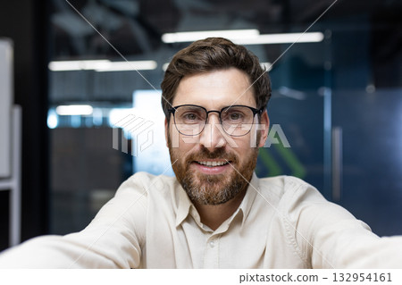 Close-up portrait of a young smiling man with glasses and a beard sitting in the office, talking and looking at the camera he is holding in his hands. Close-up portrait of a young smiling man with glasses and a beard sitting in the office, talking and looking at the camera he is holding in his hands. 132954161