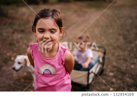 Smiling girl pulling her twin sister in a cart with a dog by her side 132954169