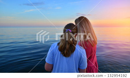 View of young mother with long bright hair in maxi dress holding hand of daughter. A walk around on wet sandy beach near wavy sea. View of young mother with long bright hair in maxi dress holding hand of daughter. A walk around on wet sandy beach near wavy sea. 132954192