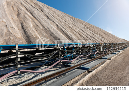 Long blue conveyor belt transports white potash ore material toward massive potassium mining dump sunlight. Industrial machinery stretches distance mineral extraction continuous production process Long blue conveyor belt transports white potash ore material toward massive potassium mining dump sunlight. Industrial machinery stretches distance mineral extraction continuous production process 132954215