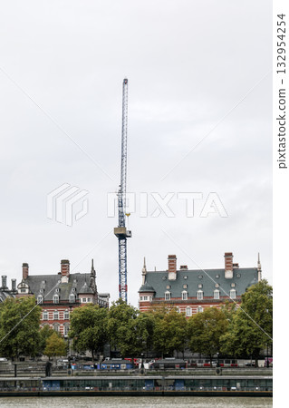Construction crane towering above historic buildings and trees by the river on a cloudy day  132954254