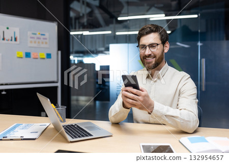 A smiling young man works in the office on a notebook, sits behind a desk on a whiteboard with stickers and uses a mobile phone in his hands. A smiling young man works in the office on a notebook, sits behind a desk on a whiteboard with stickers and uses a mobile phone in his hands. 132954657