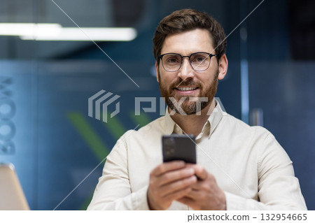 Close-up portrait of a young smiling businessman in glasses and a shirt who is in the office, holding a mobile phone and looking at the camera. Close-up portrait of a young smiling businessman in glasses and a shirt who is in the office, holding a mobile phone and looking at the camera. 132954665