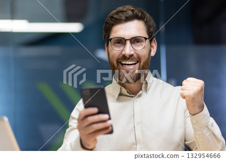 Close-up portrait of a young happy man in glasses and a light shirt who is in the office, rejoices in success and victory, holds a phone in his hand. Close-up portrait of a young happy man in glasses and a light shirt who is in the office, rejoices in success and victory, holds a phone in his hand. 132954666