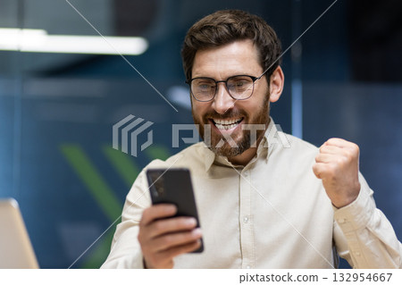 Close-up photo of a young happy man sitting in the office at the table and rejoicing in success, looking at the mobile phone screen and showing a victory gesture with his hand. Close-up photo of a young happy man sitting in the office at the table and rejoicing in success, looking at the mobile phone screen and showing a victory gesture with his hand. 132954667