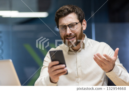 Close-up photo of an angry young businessman who is in the office, looking disappointed and upset at the screen of the mobile phone he is holding in his hand. Close-up photo of an angry young businessman who is in the office, looking disappointed and upset at the screen of the mobile phone he is holding in his hand. 132954668