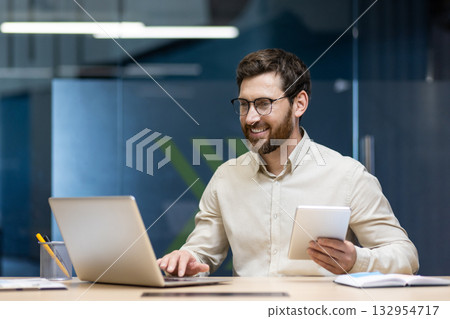 A smiling young male businessman sits in the office and works at a laptop, holding a tablet in his hands. 132954717
