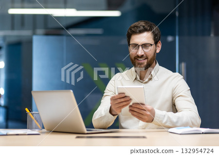 A young male office worker in glasses sits at a desk with a laptop and smilingly uses a tablet in his hands. A young male office worker in glasses sits at a desk with a laptop and smilingly uses a tablet in his hands. 132954724