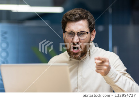 Close-up photo of a young angry man sitting in the office at the desk and shouting while pointing his finger at the laptop screen. 132954727