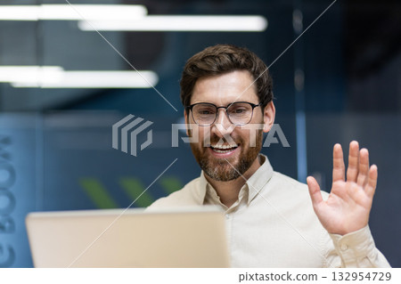 Close-up photo of a young smiling man sitting in an office at a workplace in front of a laptop, greeting and waving at the camera. Close-up photo of a young smiling man sitting in an office at a workplace in front of a laptop, greeting and waving at the camera. 132954729