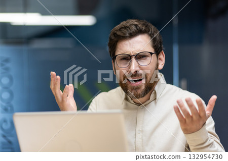 Close-up portrait of a young disappointed and sad man sitting in the office at a laptop, looking at the camera and spreading his hands. Close-up portrait of a young disappointed and sad man sitting in the office at a laptop, looking at the camera and spreading his hands. 132954730