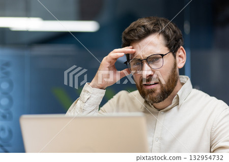 Close-up portrait of a young upset male businessman sitting in the office in front of a laptop and holding his head in frustration. Close-up portrait of a young upset male businessman sitting in the office in front of a laptop and holding his head in frustration. 132954732