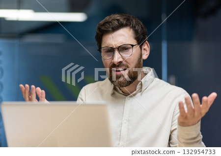 Angry young man sitting in the office with a desk, looking at the laptop screen and throwing up his hands in frustration. Angry young man sitting in the office with a desk, looking at the laptop screen and throwing up his hands in frustration. 132954733