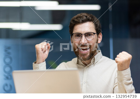A close-up portrait of a young happy and successful businessman who is in the office, sitting at the workplace and is happy to the camera, showing a victory gesture with his hands. 132954739