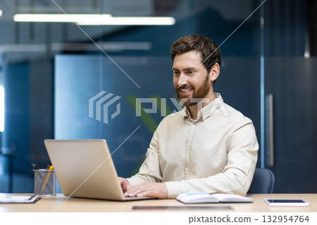 A young male businessman works in the office at a laptop, sits at a desk and looks at the screen with a smile. 132954764