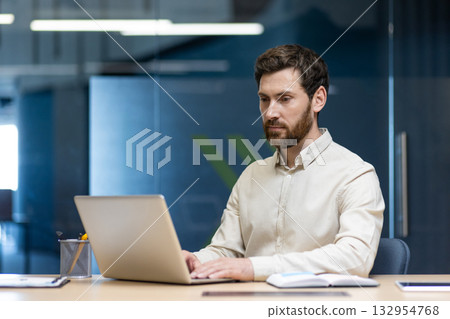 A serious and focused young man in a shirt is sitting at a desk and working on a laptop in the office. A serious and focused young man in a shirt is sitting at a desk and working on a laptop in the office. 132954768