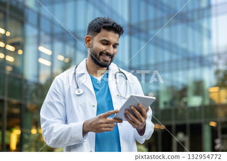A smiling medical professional wearing white coat is using a tablet outdoors, conveying professionalism, knowledge, and approachability, with a background of modern healthcare facilities. A smiling medical professional wearing white coat is using a tablet outdoors, conveying professionalism, knowledge, and approachability, with a background of modern healthcare facilities. 132954772