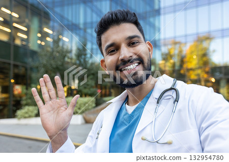 Portrait of a smiling indian doctor waving hand in front of a modern hospital building, radiating friendliness and approachability while showcasing confidence and professionalism 132954780