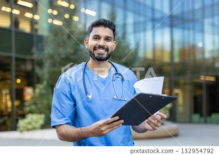 Confident and friendly indian doctor is standing outside a modern hospital building, holding a clipboard with patient medical records and smiling Confident and friendly indian doctor is standing outside a modern hospital building, holding a clipboard with patient medical records and smiling 132954792