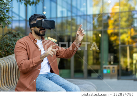 Young man is interacting with virtual reality using a vr headset outdoors, exploring immersive technology against a backdrop of modern architecture 132954795