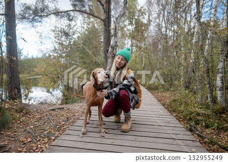 Woman hiker on daily walk in autumn forest together with dog, sitting on wooden trail near lake. Woman hiker on daily walk in autumn forest together with dog, sitting on wooden trail near lake. 132955249
