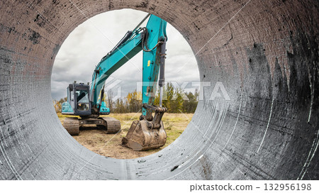 Through the circular opening of a pipe, a construction excavator works diligently at the site under overcast skies, preparing the ground for new projects 132956198