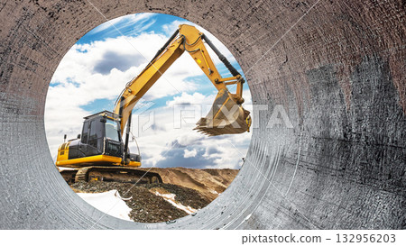 Heavy machinery operates at a construction site as seen through a circular pipe. The excavator skillfully raises its bucket against a backdrop of clouds and earth 132956203