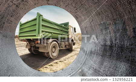 A construction site reveals a green truck parked on rough terrain, framed by a large pipe, with a blue sky stretching above and construction activities in motion A construction site reveals a green truck parked on rough terrain, framed by a large pipe, with a blue sky stretching above and construction activities in motion 132956221