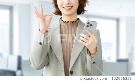 A smiling woman/businesswoman holding a smartphone and making an OK pose 132956331
