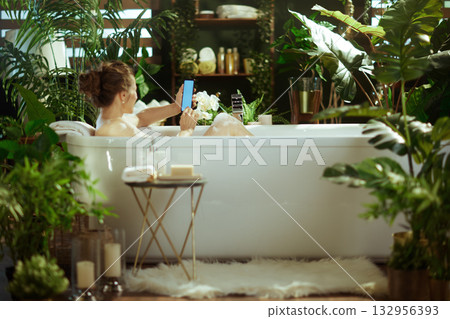 Woman Relaxing in Bathtub with Phone Amidst Plants 132956393