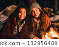 Two happy girls in winter clothes sit by a campfire in a winter forest at night. Friends having fun together, camping trip, winter outdoor recreation with family 132956489