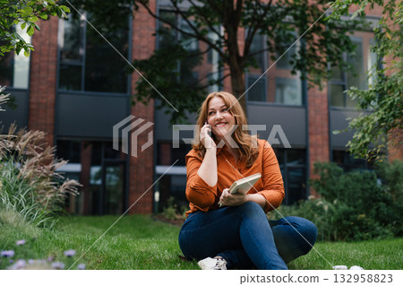 Confident woman rmaking phone call while reading book outdoors in park. Confident woman rmaking phone call while reading book outdoors in park. 132958823