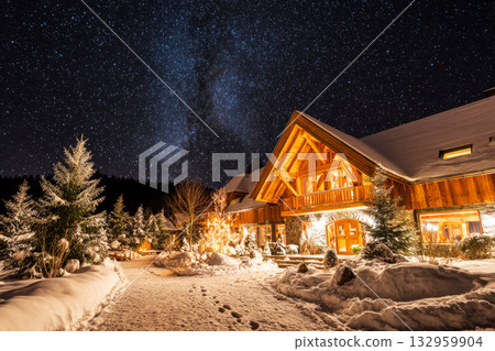 A beautiful mountain chalet stands aglow with warm lights at night, surrounded by snow-covered trees and a peaceful winter landscape. A pathway leads through the snow 132959904