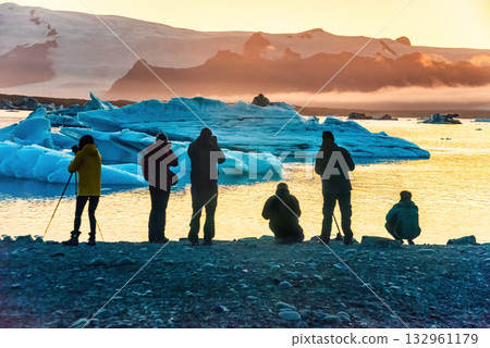 Tourists admire the stunning sunset over Iceland's Glacier Lagoon, surrounded by large icebergs. The serene scene captures the beauty of nature 132961179