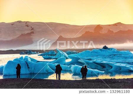 Tourists admire the stunning sunset over Iceland's Glacier Lagoon, surrounded by large icebergs. The serene scene captures the beauty of nature 132961180